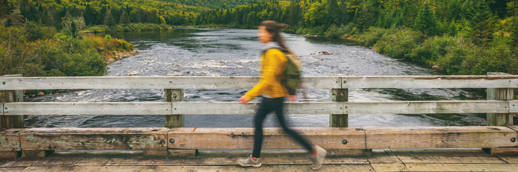 Frau mit gelber Jacke und Rucksack geht über eine hölzerne Brücke vor einem Fluss und Wald