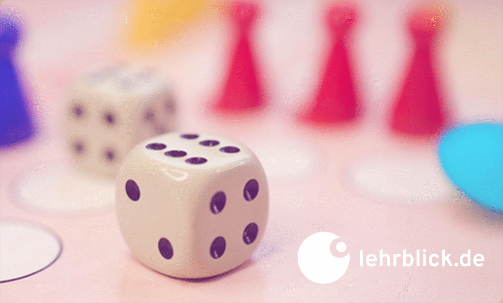 Nahaufnahme eines weißen Würfels mit schwarzen Punkten auf einem Spielbrett, im Hintergrund rote Spielfiguren und ein weiterer Würfel, unten rechts das Logo von lehrblick.de