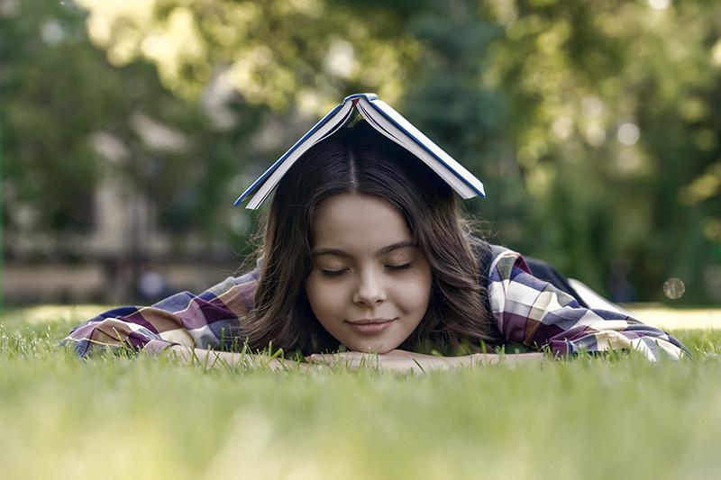 Ein Mädchen liegt in der Wiese und entspannt. Es hat ein aufgeklapptes Buch auf dem Kopf.
